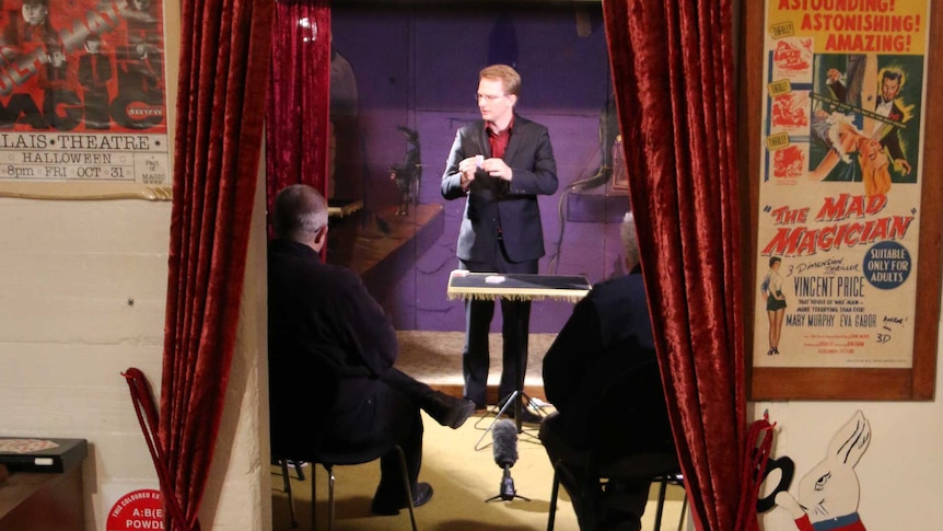 A man stands up showcasing magic trick to two men seated in front of him.