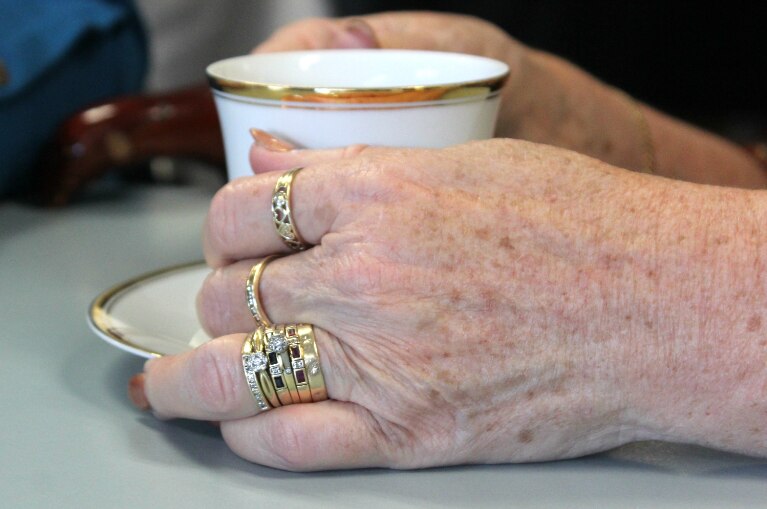 Older woman's hand with eight gold rings holds gold-edged tea cup.