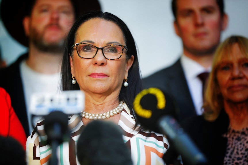 A middle-aged Indigenous woman with long dark hair stands behind a lectern filled with microphones.