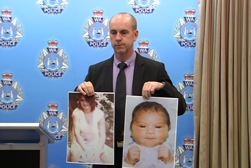 A middle-aged man in a black suit and purple shirt holds up a photo of a woman and a baby. Police backdrop behind him.