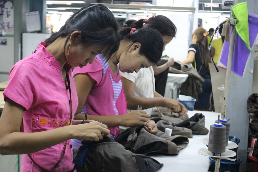Three women wearing pink tops stand in a row and sew brown garments by hand.