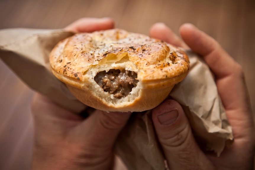 Close-up photo of two hands holding a pie in a brown paper bag. The pie has a small bite at the front, revealing meat inside.
