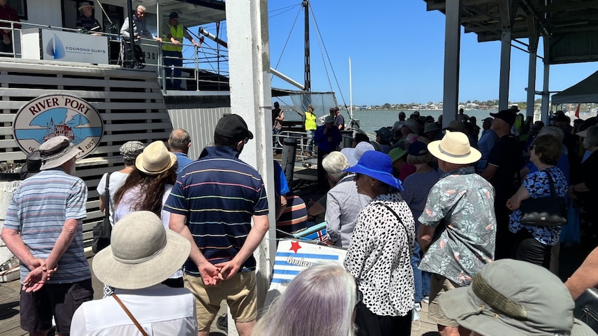 A group of people stand on the wooden wharf as several men address them from aboard a paddle steamer boat