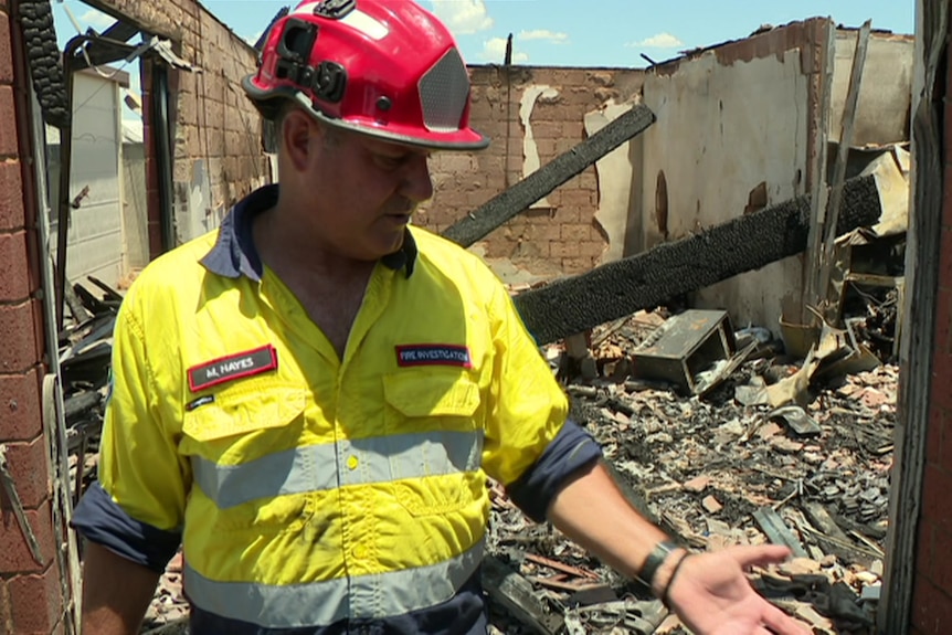 A man in high-vis gear and a red helmet stand in a burned building. 