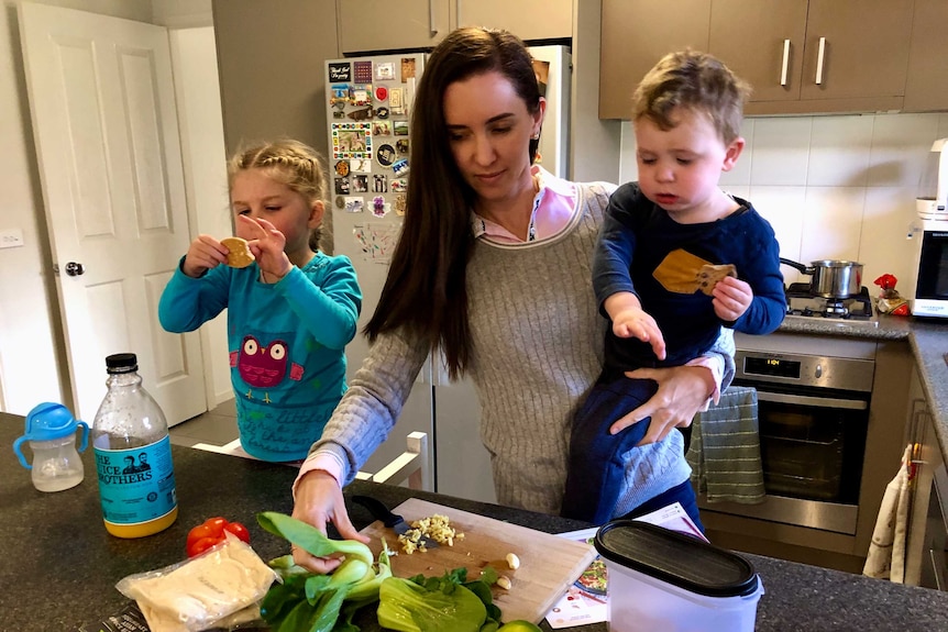 A woman stands over a kitchen counter, preparing dinner while holding a toddler. Her younger daughter stands beside her.