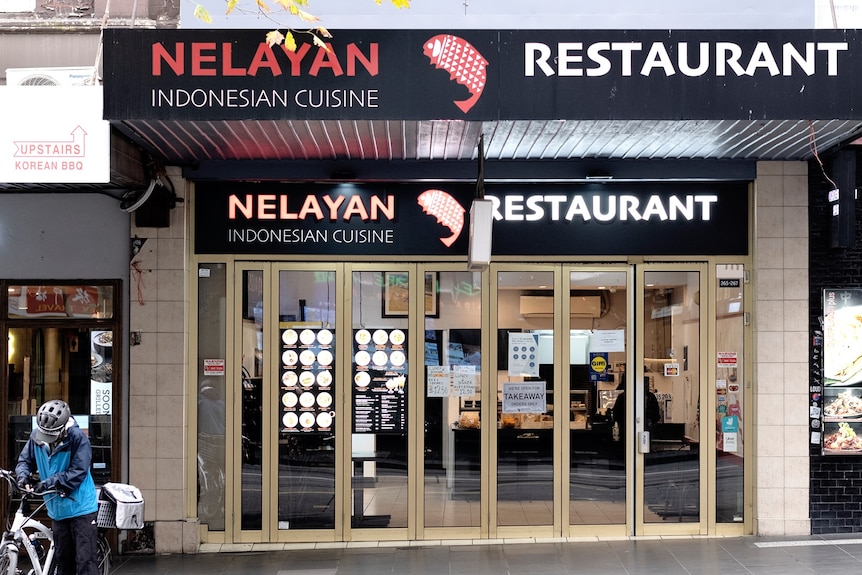 An exterior building of a restaurant with a delivery man holding a bike standing in the corner 