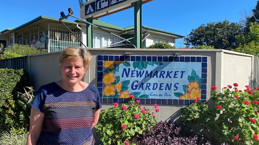 A smiling woman stands outside a caravan park sign that says 'Newmarket gardens'