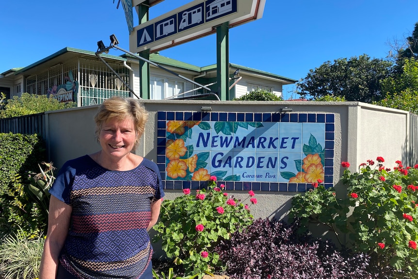 A smiling woman stands outside a caravan park sign that says 'Newmarket gardens'