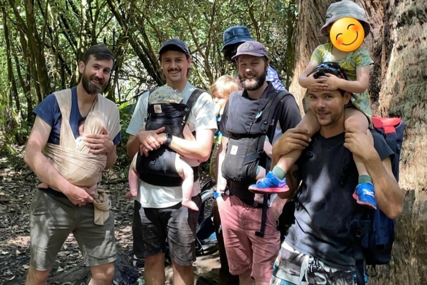 Five men in a bush setting either carry a baby in front pack or a toddler on their shoulders