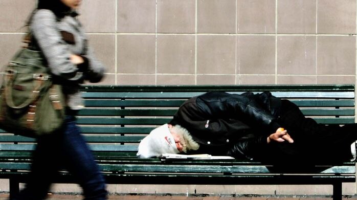 picture of a homeless man on a bench as a passerby strides past.