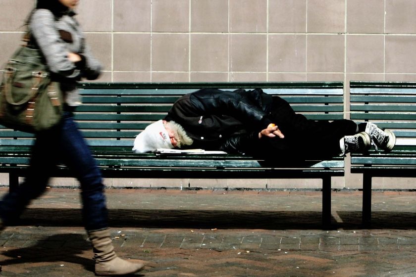 A homeless man sleeps on a bench in Sydney's CBD as a woman walks past him.