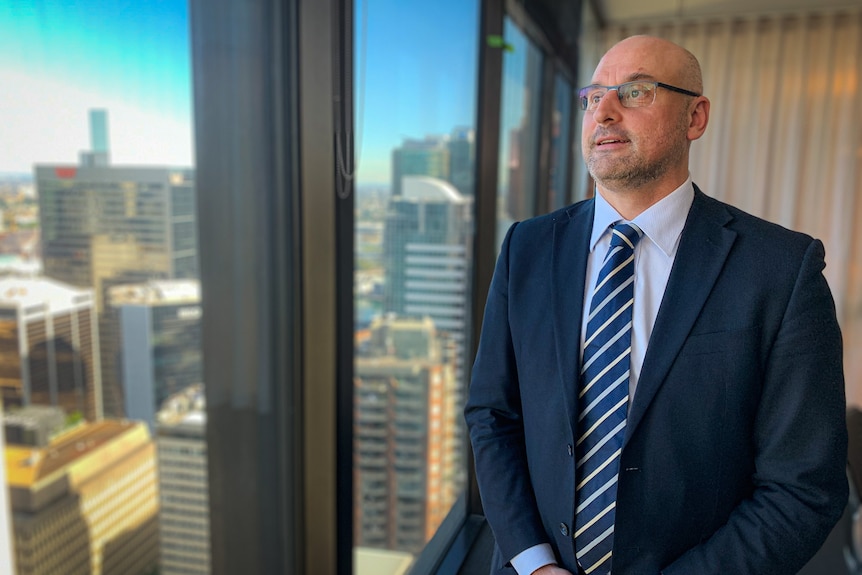 A broad-shouldered balding man with glasses smiles as he looks out to his right over other high-rise office buildings.