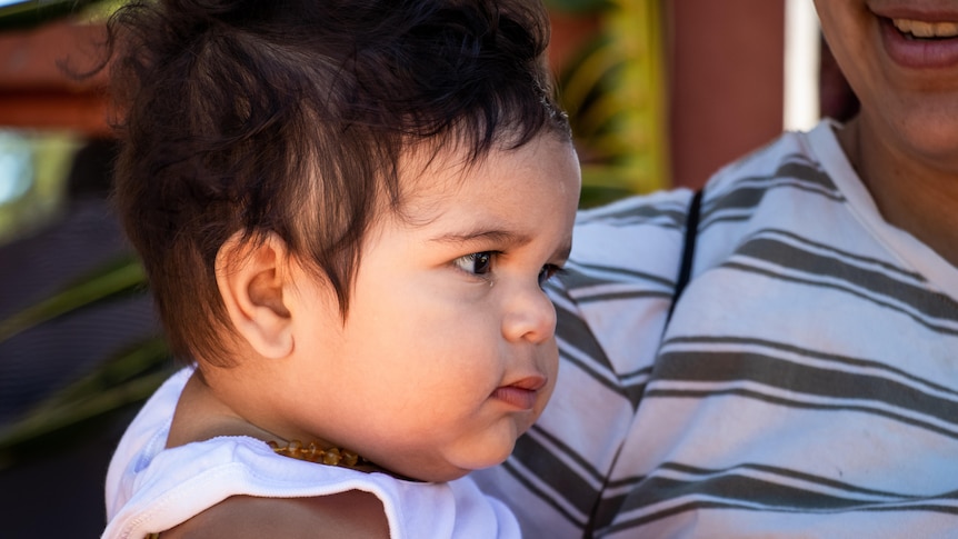 A baby in a white shirt smiles and looks off camera.