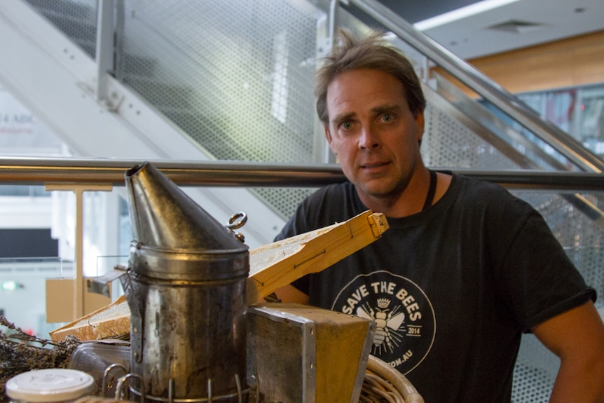 A man wearing a t-shirt reading 'save the bees' sits next to a basket of beekeeping equipment.