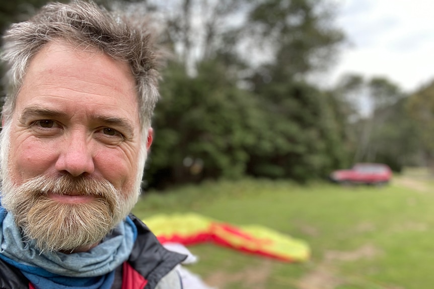 Man in beard smiles at camera to the left of frame, blurred vegetation behind him.