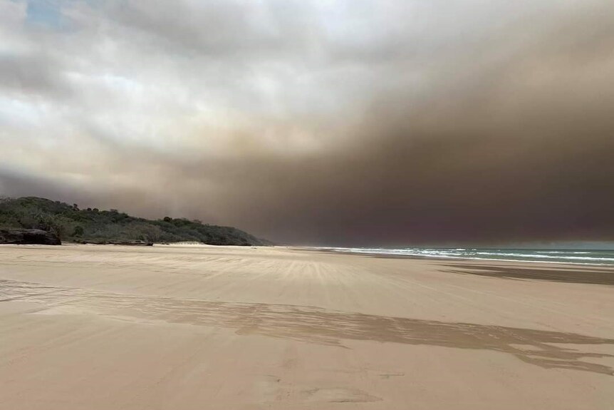 A beach is seen with a grassy hilll to the left and the sea to the right, with smoke in the sky above.