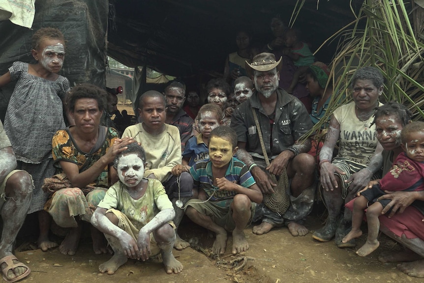 A large family, some with paint decorating their bodies, crouch in front of an erected tarpaulin