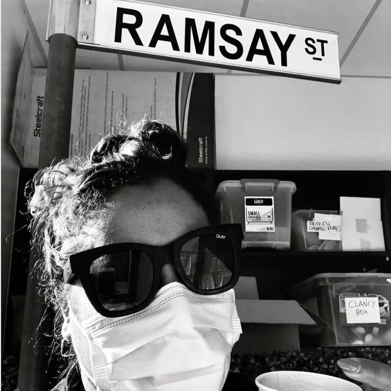 Shareena Clanton wears a face mask in a black-and-white photo of her underneath a 'Ramsay St' sign.