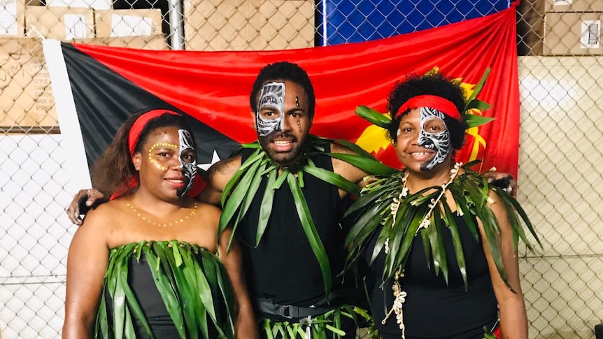 A man stands in between two women wearing grass skirts and attire. All three stand in from of a PNG flag