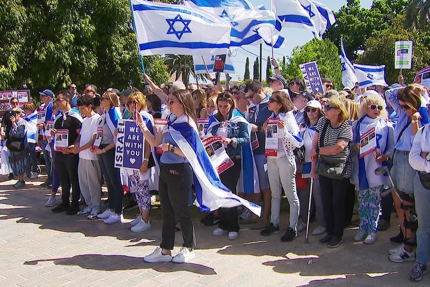 A crowd of people, many draped in Israeli flags, at a rally on a sunny day.