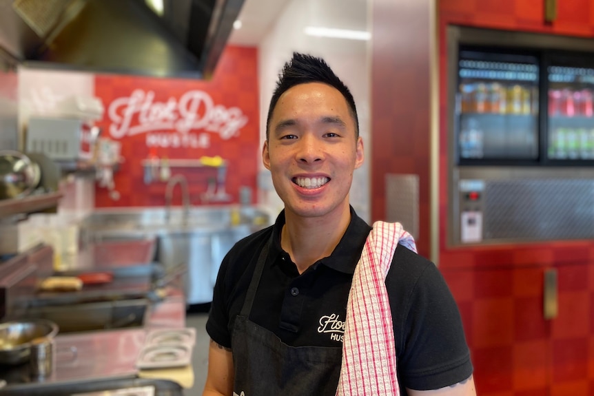 Man with tea towel over shoulder stands in front of commercial kitchen