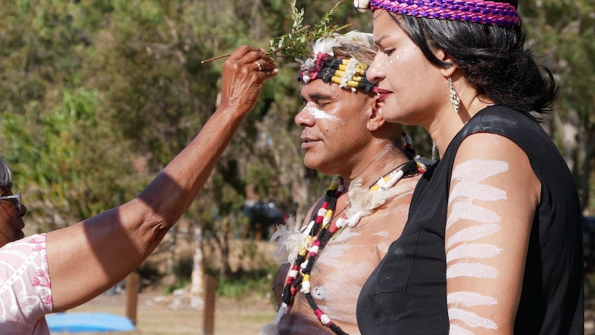 A man and woman stand with their eyes closed while a woman brushes their face with a tree branch.