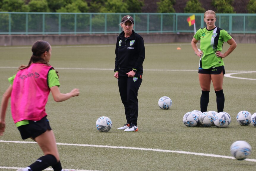 A female soccer coach stands among a bunch of soccer balls.