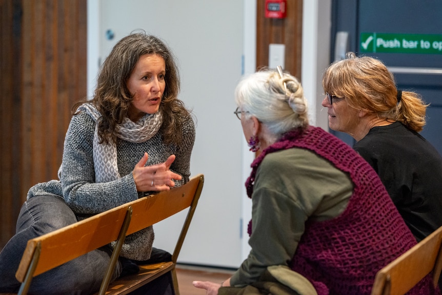 Caroline leans over chairs to talk to two older women. 