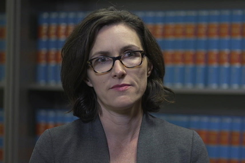 A woman wearing a grey blazer and tortoise-shell spectacles looks to the right of the camera.