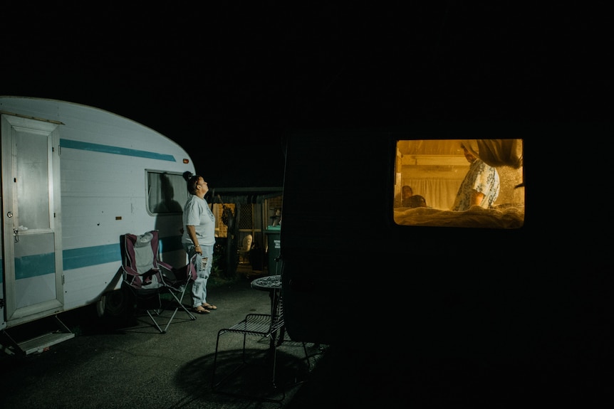 Binny stands in a space with chairs between two caravans at night. Her son can be seen through the window of one of the vans.