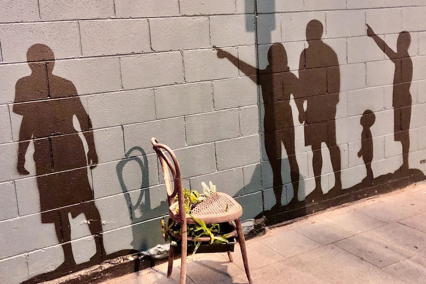 A cane chair in front of a gray brick wall with five people's shadows on it