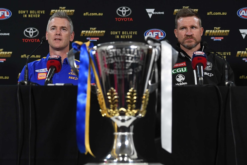 Two men sit at a table with a large, silver cup in the foreground.