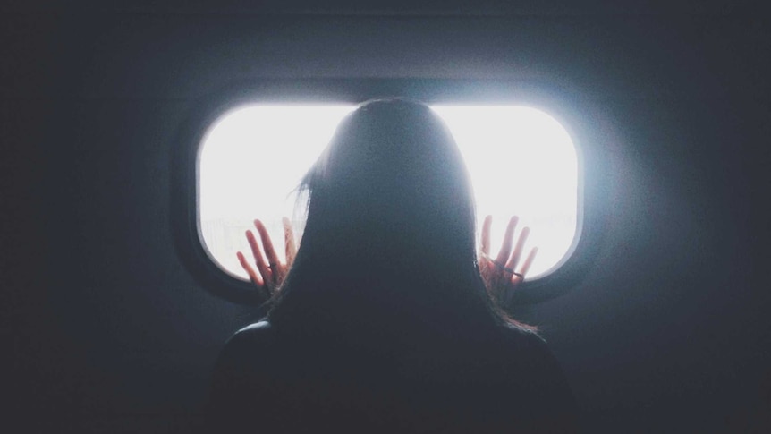 A woman stands facing at a window in a darkened room, her hands on the glass.