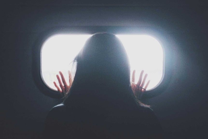A woman stands facing at a window in a darkened room, her hands on the glass.