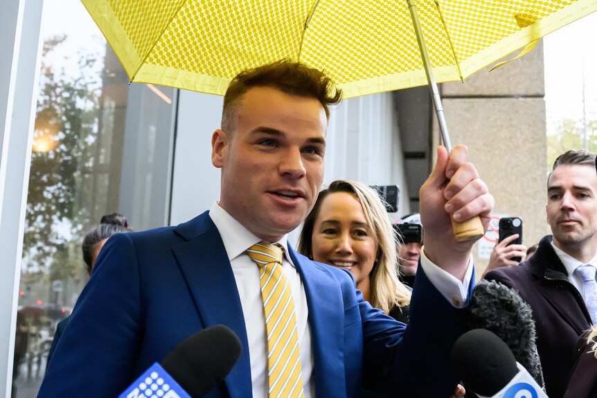 A man in a black suit leaves a court building, sheltered by a bright yellow umbrella and followed by members of the media.