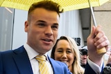 A man in a black suit leaves a court building, sheltered by a bright yellow umbrella and followed by members of the media.