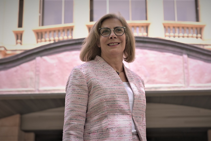 A woman with blonde hair wearing a pink suit jacket and black glasses stands in front of a court building