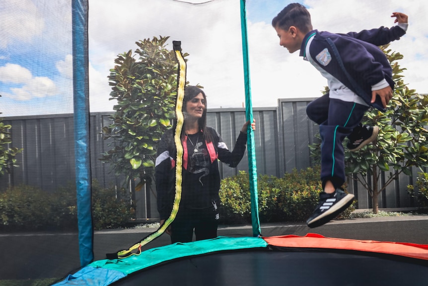 A woman stands outside a safe trampoline as her son bounces up and down. Both are wearing tracksuits