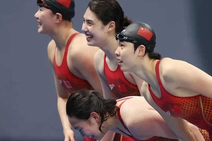  四个穿着红色和黑色套件的女游泳运动员在查看成绩板时微笑着向前弯腰。