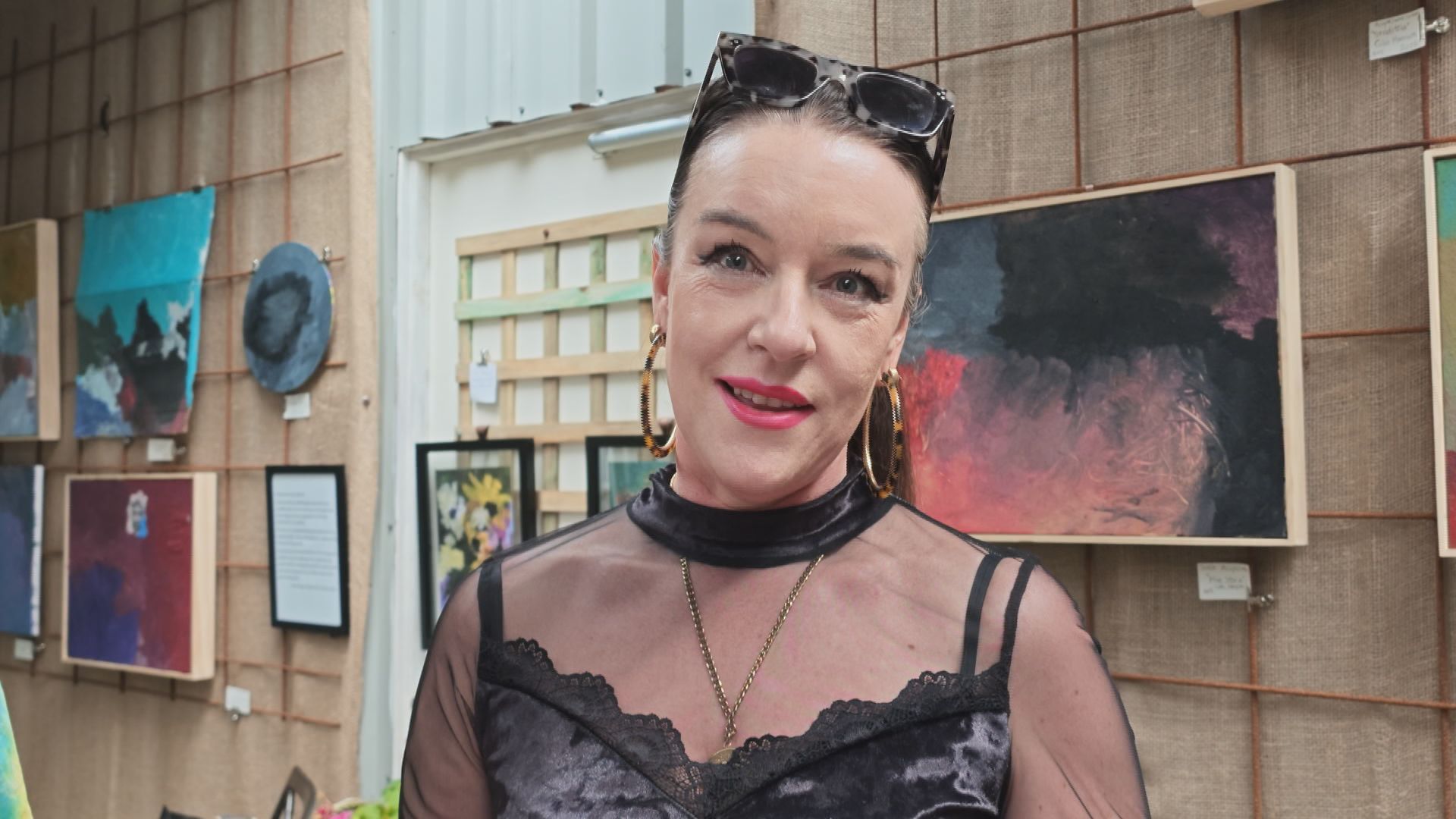 Serena McLaughlan stands with artwork on the wall behind her. She wears black, lacy clothes and large gold hoop earrings.