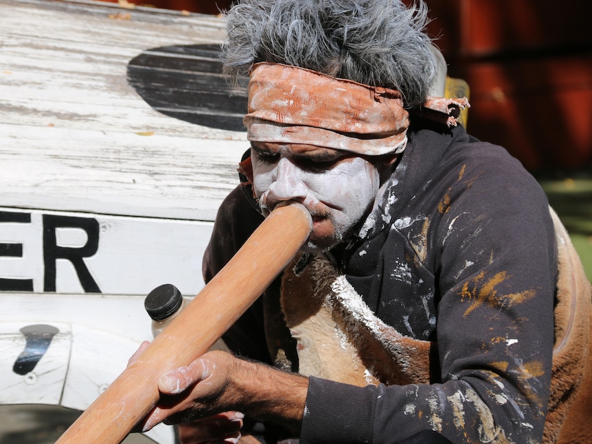 An Indigenous man, wearing traditional face paint, plays the didgeridoo.