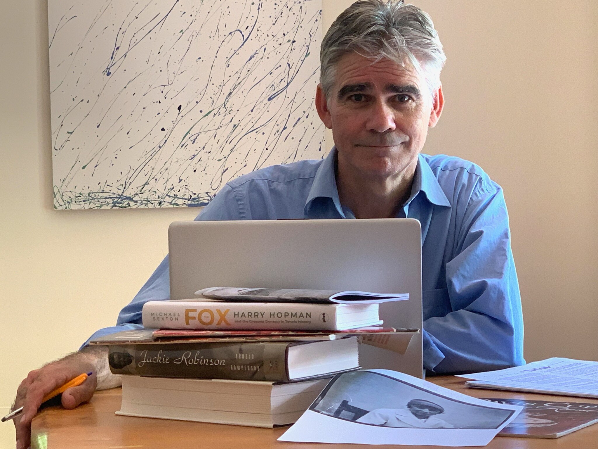 A man in a blue shirt and gray hair looks at the camera while sitting behind an open laptop and a stack of books.