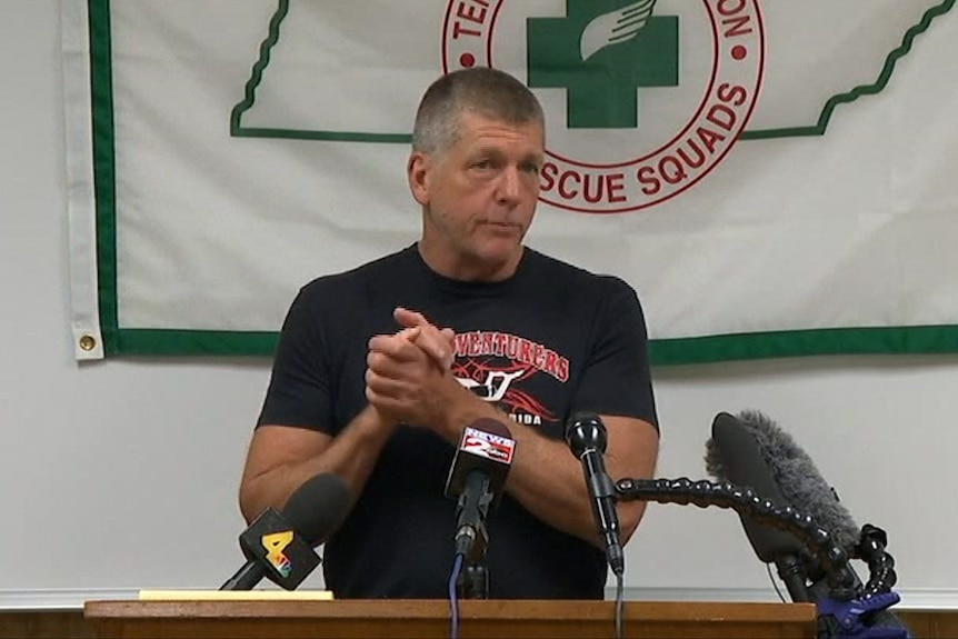 A man in a t-shirt addresses reporters with several microphone placed in front of him.