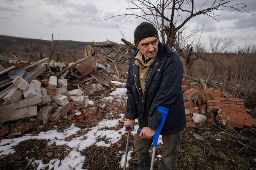 Andrii Cherednichenko on crutches walks next to ruins in Kamyanka, Ukraine