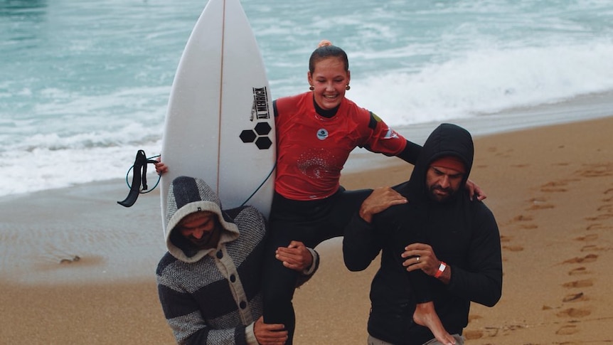 A girl with a surfboard is carried onshore by two men.