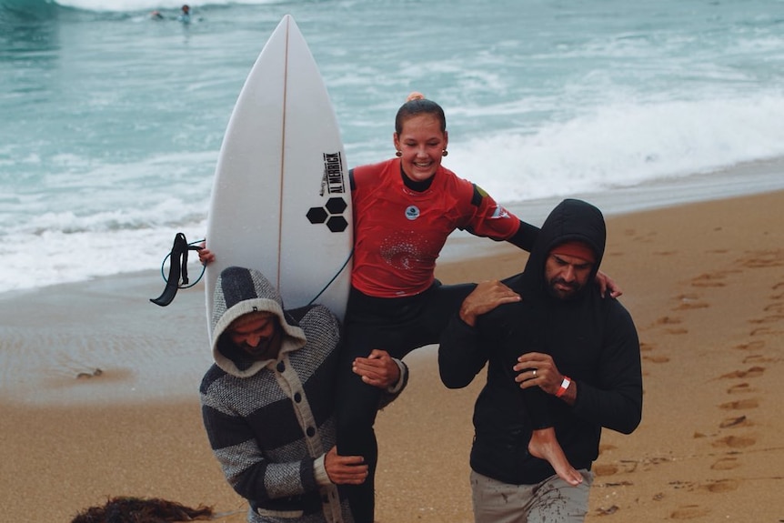 A girl with a surfboard is carried onshore by two men.