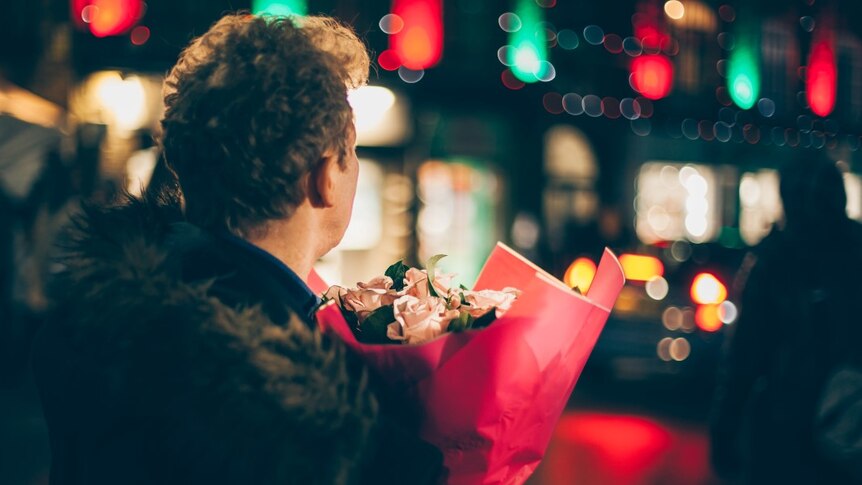 Man looks away from the camera, whilst holding a bunch of flowers in the city at night time.