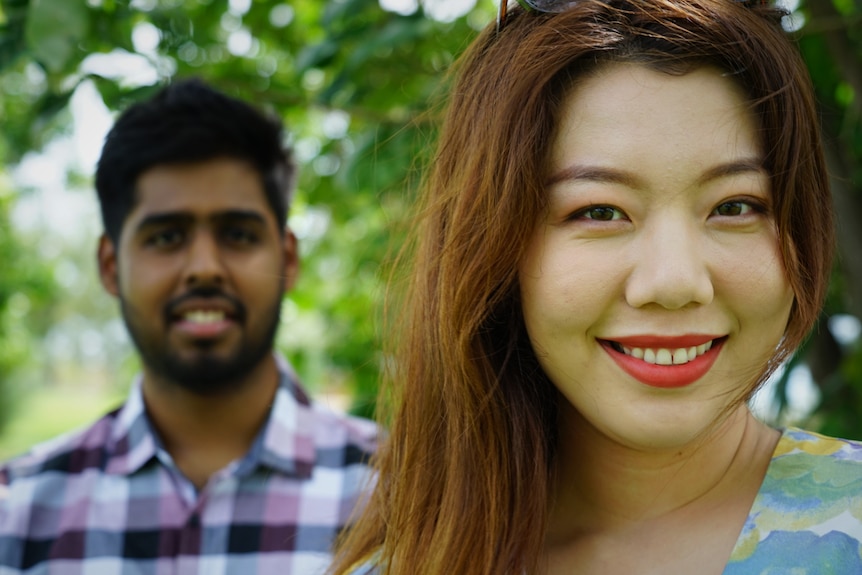 A young woman and man stand facing a camera outside with green tress behind them