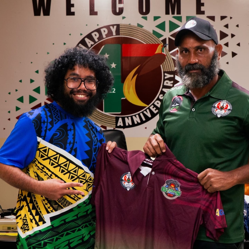 A reporter and the governor stand smiling in an office holding a PNG kumuls rugby jersey