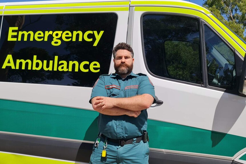 A white man with brown hair and a beard with his arms crossed in front of an ambulance.